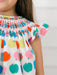 Close-up of a child wearing a white smocked dress with colorful apple print and ruffled sleeves.