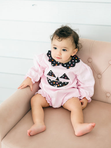 A baby girl sitting on a dusty pink tufted armchair, wearing a light pink long-sleeve romper with a black polka-dot collar and a jack-o'-lantern face applique on the chest.