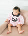 A baby girl sitting on a dusty pink tufted armchair, wearing a light pink long-sleeve romper with a black polka-dot collar and a jack-o'-lantern face applique on the chest.