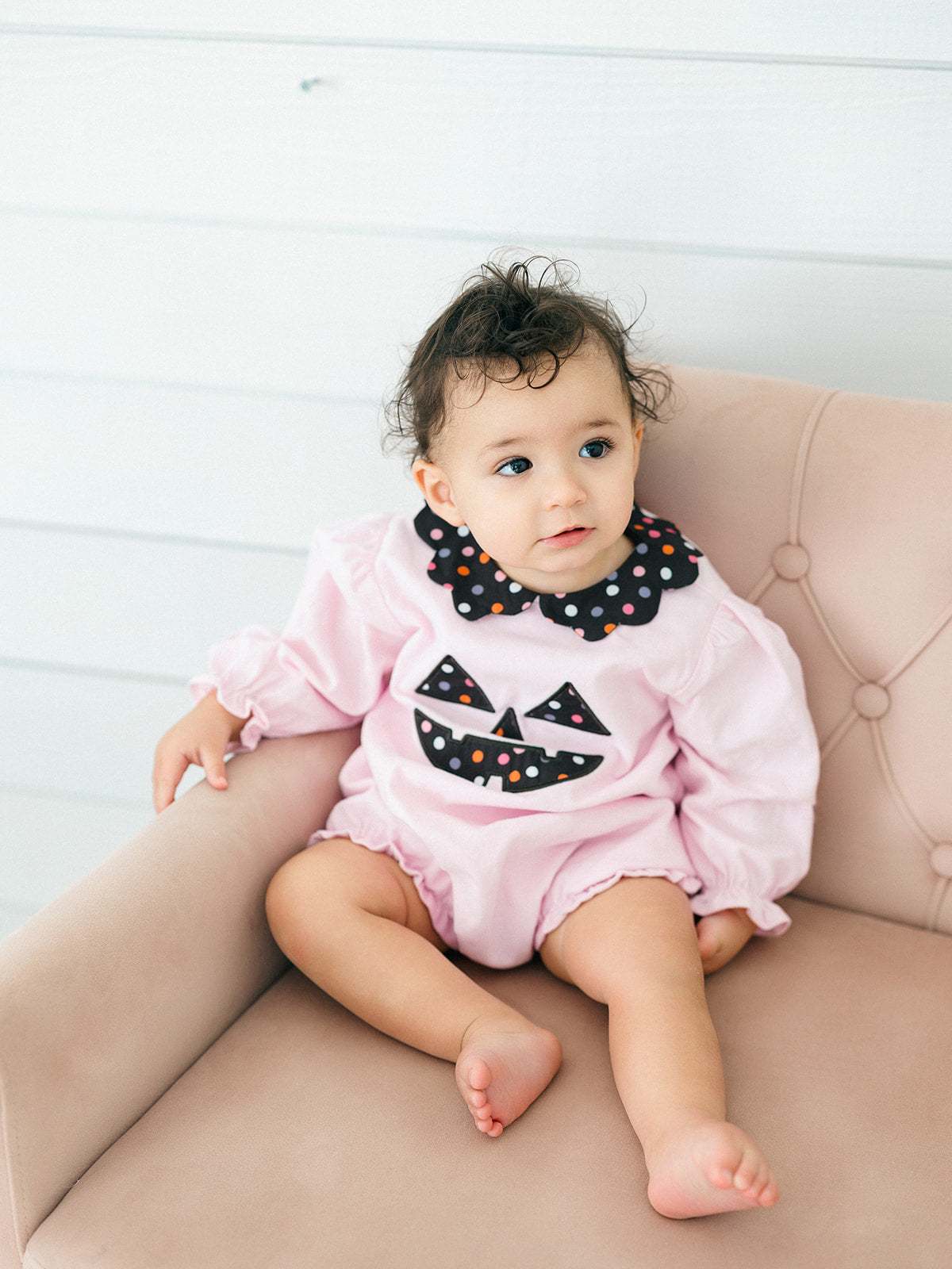 A baby girl sitting on a dusty pink tufted armchair, wearing a light pink long-sleeve romper with a black polka-dot Peter Pan collar and a black jack-o'-lantern face applique on the front.