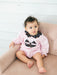 A baby girl sitting on a dusty pink tufted armchair, wearing a light pink long-sleeve romper with a black polka-dot Peter Pan collar and a black jack-o'-lantern face applique on the front.