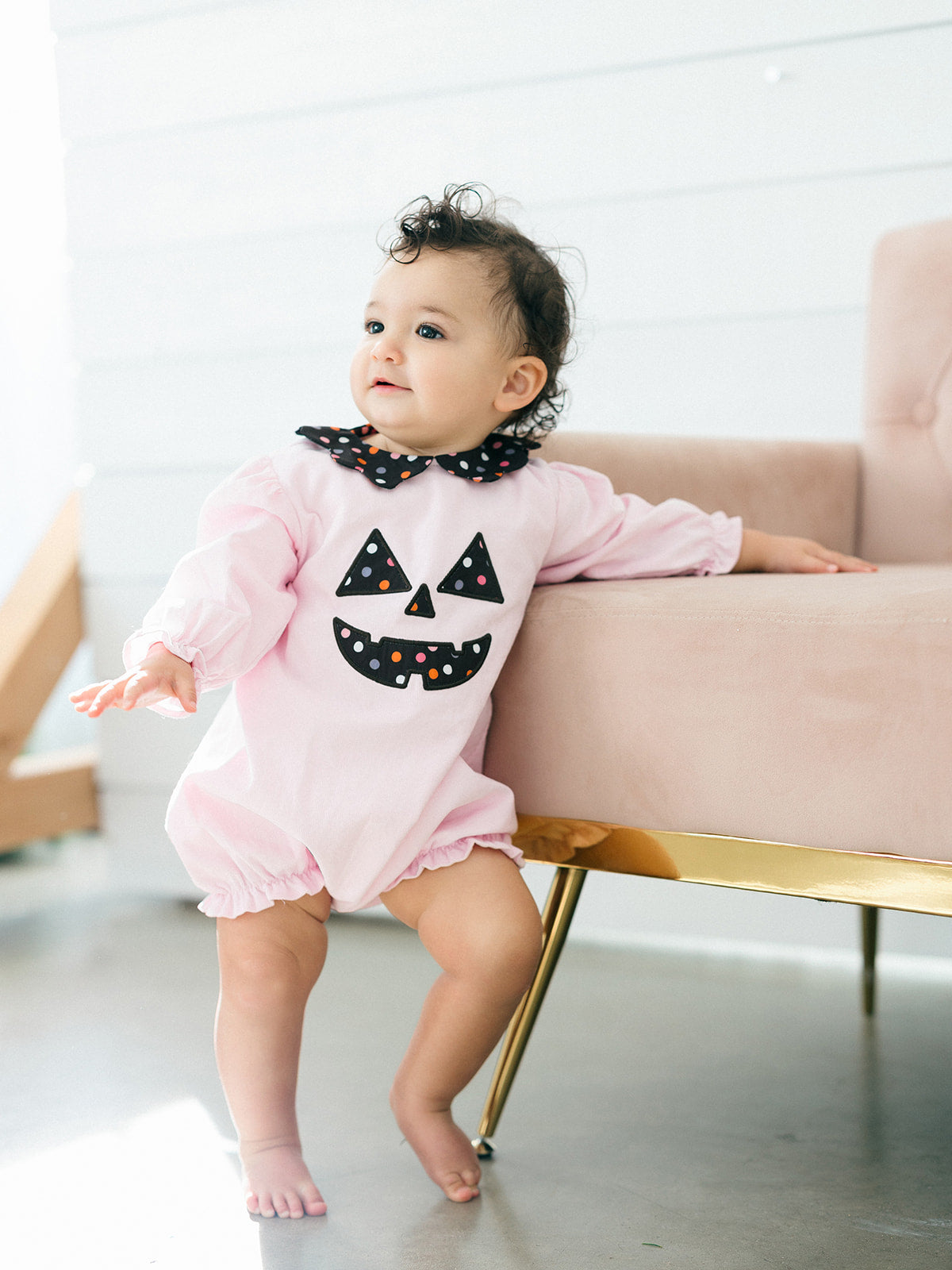 A baby wearing a light pink Halloween jumpsuit with a black polka-dot Peter Pan collar and a black jack-o'-lantern face on the front, standing next to a pink armchair with gold legs.