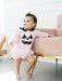 A baby wearing a light pink Halloween jumpsuit with a black polka-dot Peter Pan collar and a black jack-o'-lantern face on the front, standing next to a pink armchair with gold legs.