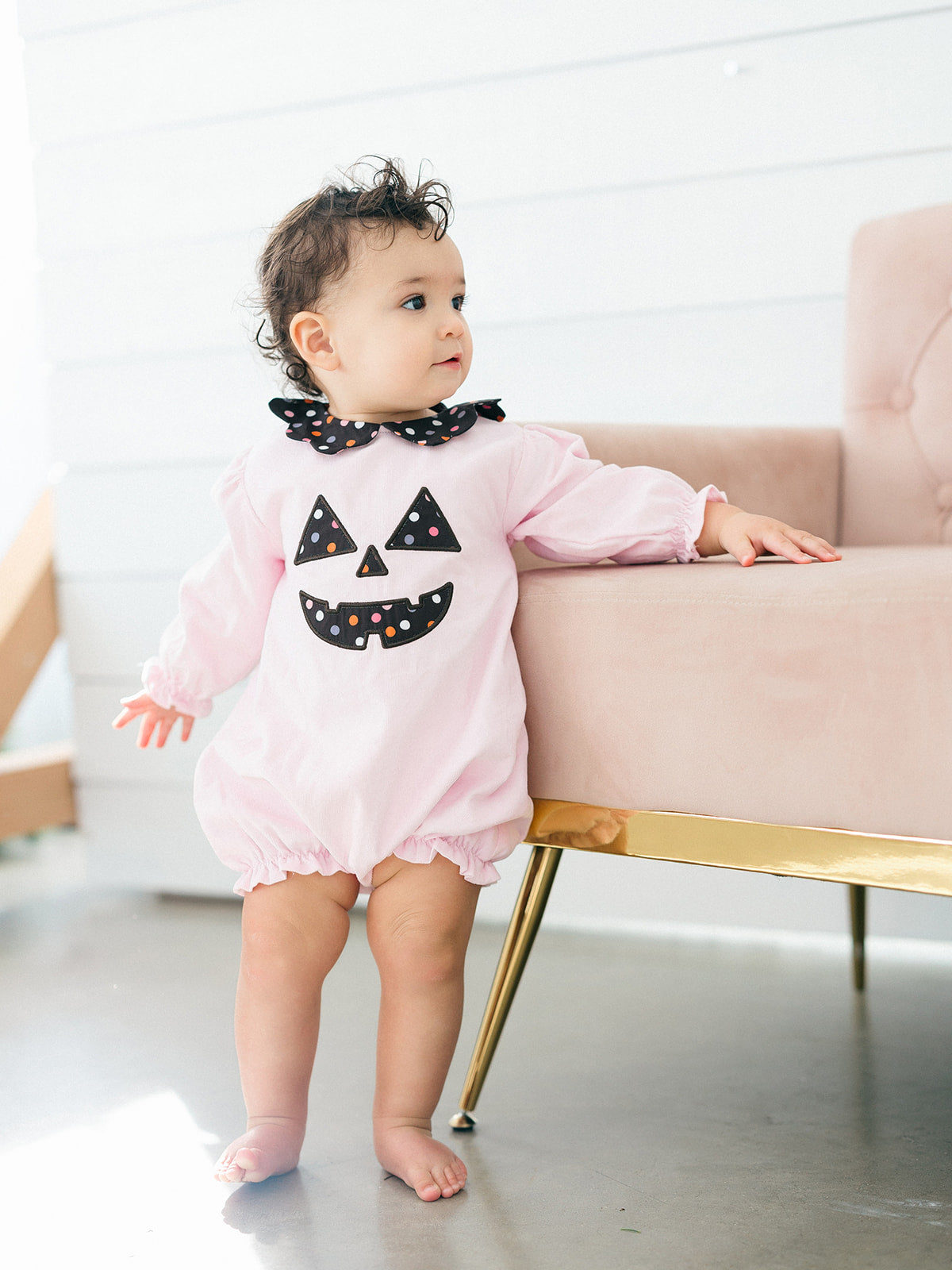A baby wearing a light pink long-sleeve romper with a black polka-dot Peter Pan collar and a black jack-o'-lantern face applique on the chest, standing next to a pink sofa with gold legs.