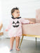 A baby wearing a light pink long-sleeve romper with a black polka-dot Peter Pan collar and a black jack-o'-lantern face applique on the chest, standing next to a pink sofa with gold legs.