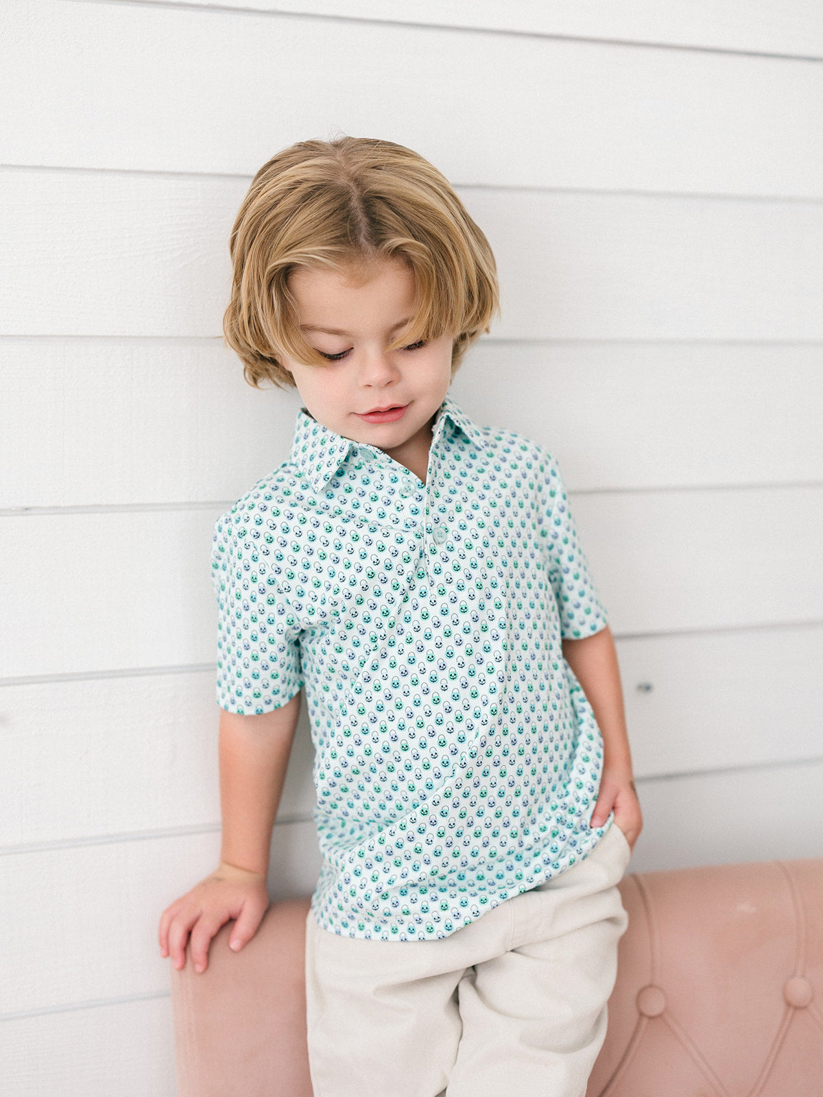 A young boy with blonde hair wearing a light blue and white pumpkin faces patterned short-sleeve button-up shirt and beige pants, leaning against a white paneled wall.