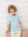 A young boy with blonde hair wearing a light blue and white pumpkin faces patterned short-sleeve button-up shirt and beige pants, leaning against a white paneled wall.