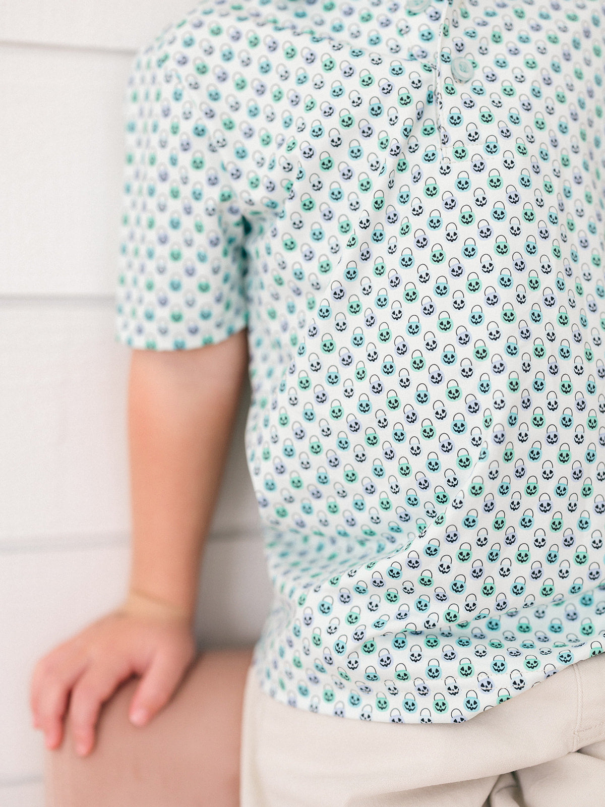 A close-up of a child wearing a white short-sleeve polo shirt with a repeating pattern of small green and black pumpkin faces, paired with light beige shorts against a white paneled wall.