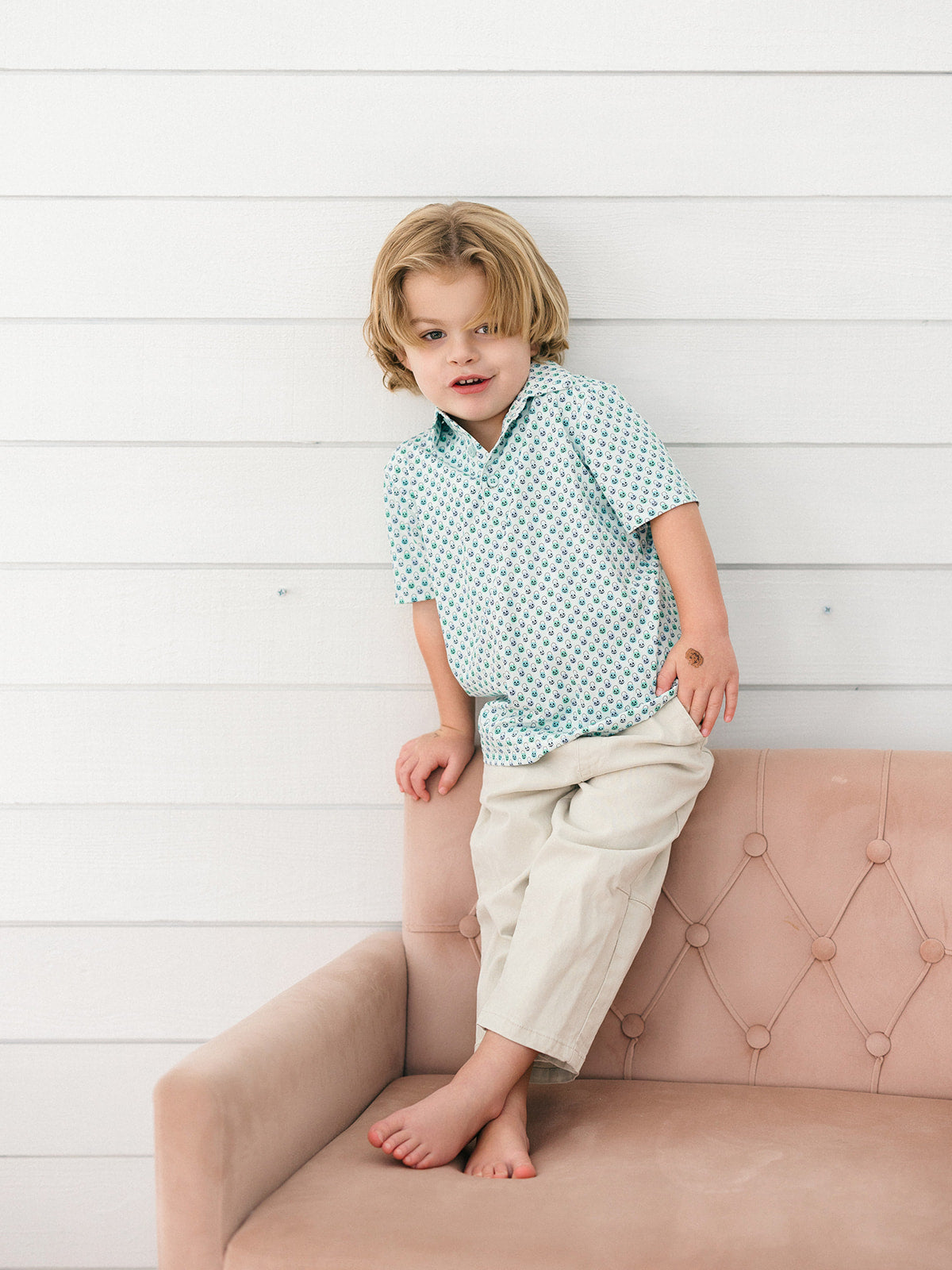 A young boy with blonde hair sits on a dusty pink tufted sofa against a white wood-paneled wall. He is wearing a light blue short-sleeve button-up shirt with a small pumpkin basket pattern and khaki colored pants.