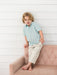 A young boy with blonde hair sits on a dusty pink tufted sofa against a white wood-paneled wall. He is wearing a light blue short-sleeve button-up shirt with a small pumpkin basket pattern and khaki colored pants.