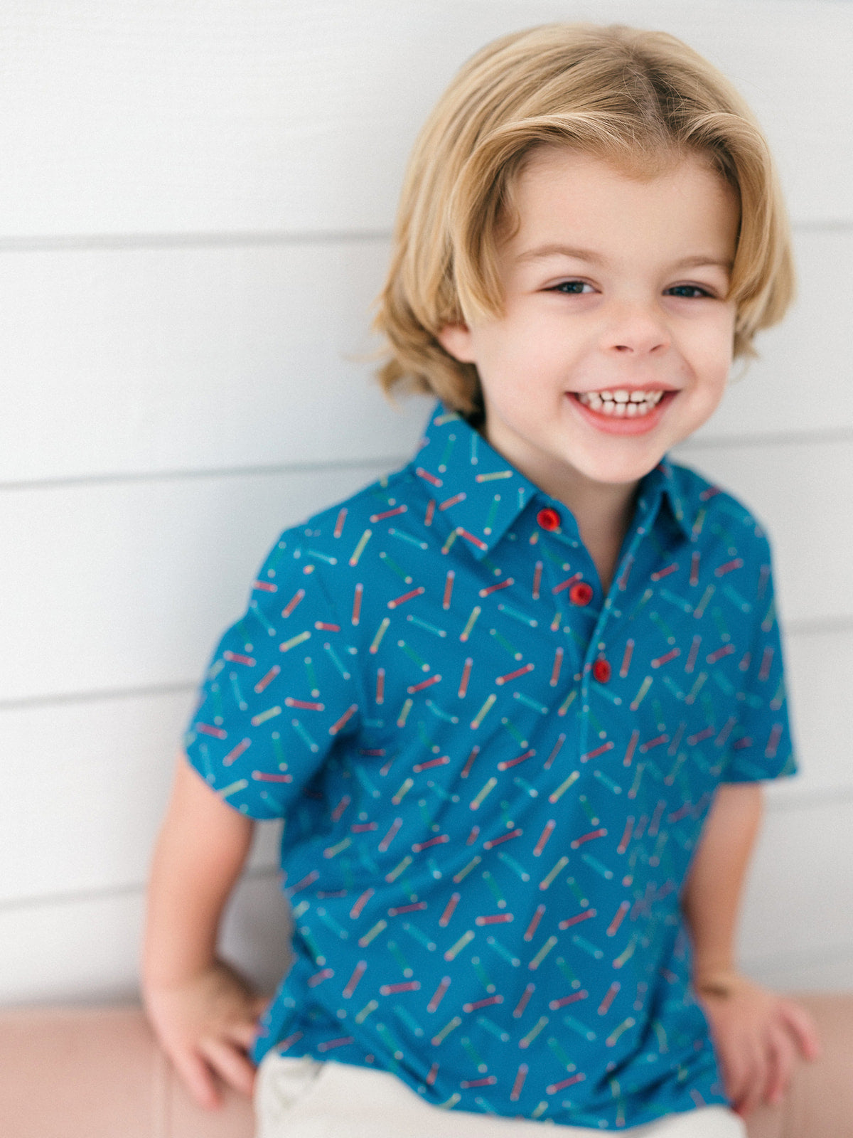 A young boy with blonde hair smiling while wearing a blue short-sleeve button-up shirt with a red, white, and blue pencil print, set against a white paneled wall.