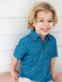 A young boy with blonde hair smiling while wearing a blue short-sleeve button-up shirt with a red, white, and blue pencil print, set against a white paneled wall.
