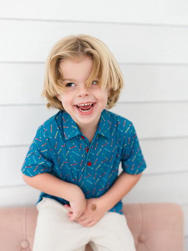A young blonde boy smiling and laughing while wearing a blue short-sleeve button-up shirt with a colorful confetti pattern and white pants, sitting on a pink tufted bench against a white paneled wall.
