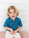 A young blonde boy smiling and laughing while wearing a blue short-sleeve button-up shirt with a colorful confetti pattern and white pants, sitting on a pink tufted bench against a white paneled wall.
