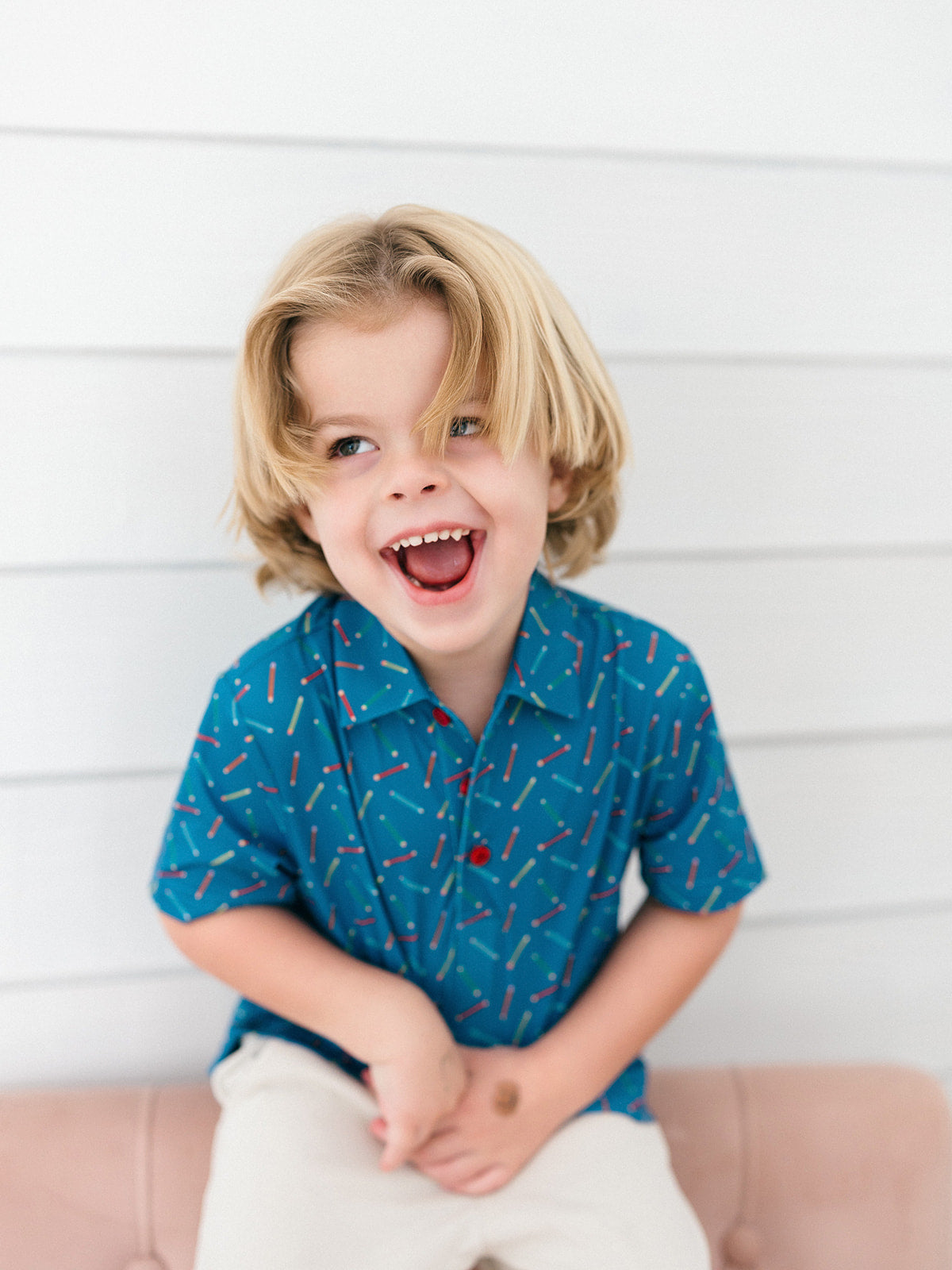 A young blonde boy wearing a blue short-sleeve button-up shirt with a red and white pencil pattern, sitting against a white shiplap wall.
