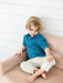 A young blonde boy sitting on a dusty pink tufted armchair, wearing a blue short-sleeve button-up shirt with a small white and red pattern, paired with cream-colored pants.