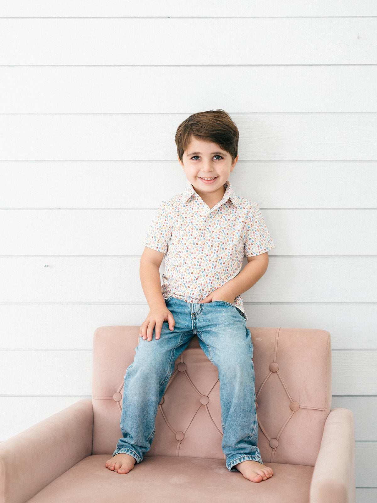 A young boy with brown hair sits barefoot on a dusty pink tufted armchair, wearing a short-sleeve button-up shirt with a tiny red, blue, and yellow pattern on a white background, paired with blue jeans, against a white shiplap wall.