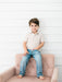 A young boy with brown hair sits barefoot on a dusty pink tufted armchair, wearing a short-sleeve button-up shirt with a tiny red, blue, and yellow pattern on a white background, paired with blue jeans, against a white shiplap wall.