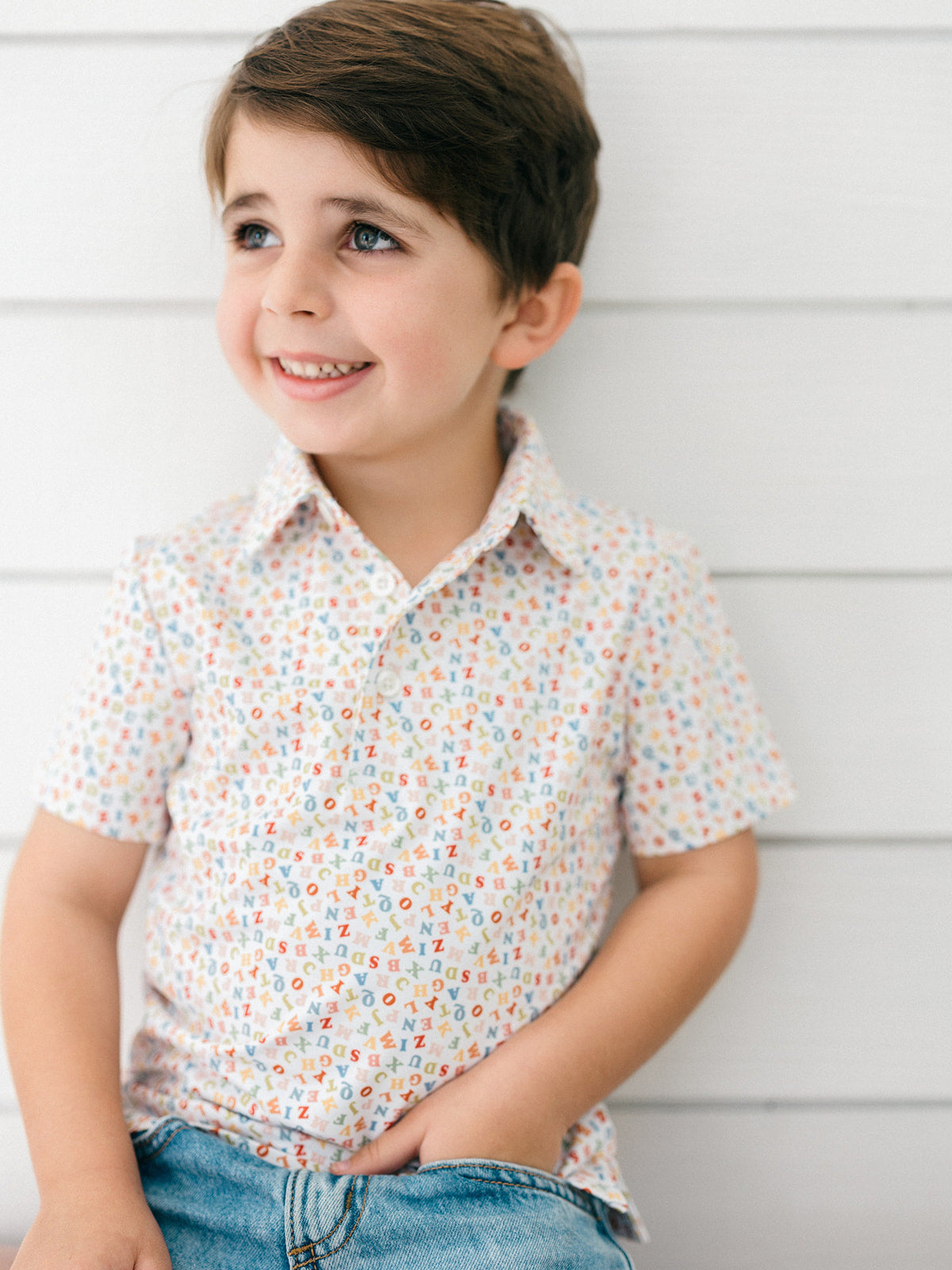 A young boy with brown hair smiling and looking to the side, wearing a short-sleeve button-up shirt with a small multicolor geometric pattern on a white base, paired with blue jeans, set against a white paneled wall.