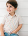 A young boy with brown hair smiling and looking to the side, wearing a short-sleeve button-up shirt with a small multicolor geometric pattern on a white base, paired with blue jeans, set against a white paneled wall.