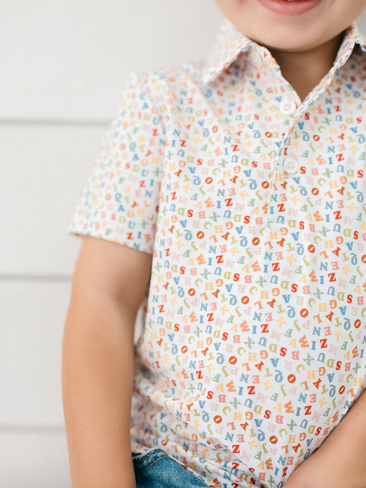 A close-up of a child wearing a short-sleeve button-up shirt with an all-over multicolor alphabet print on a white base, paired with blue jeans.