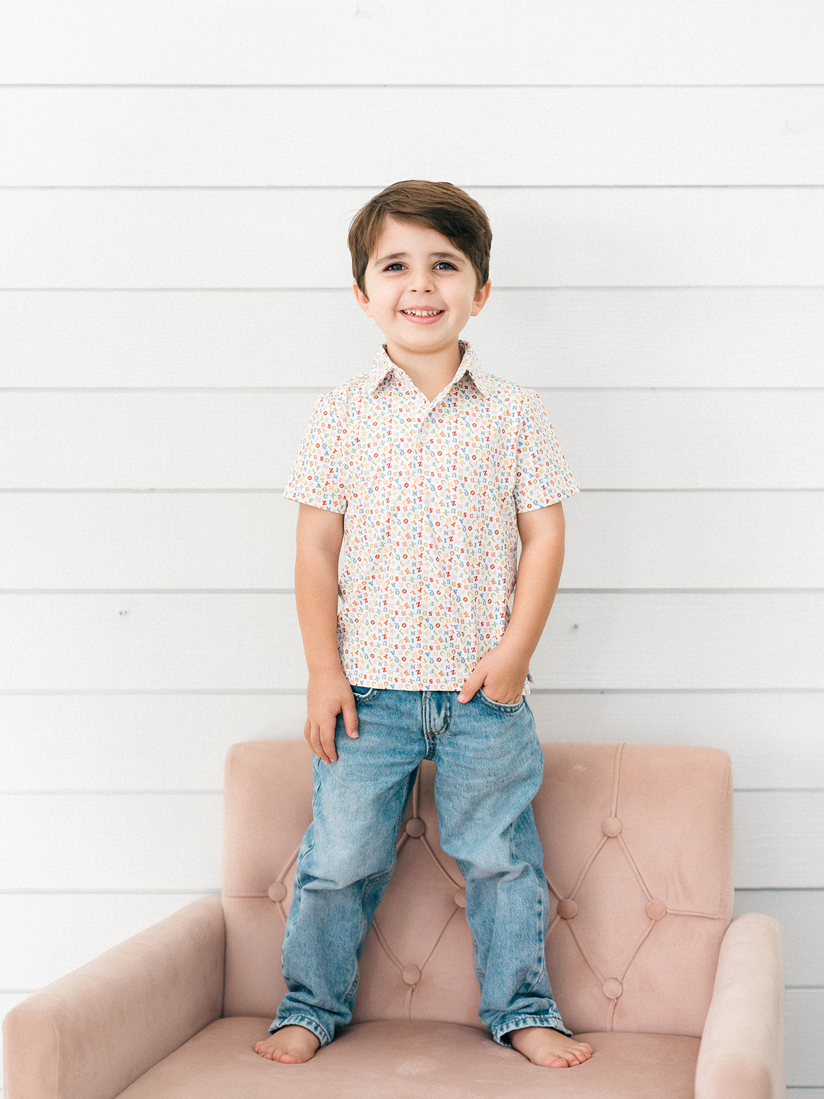 A young boy stands barefoot on a dusty pink tufted armchair against a white wood-paneled wall, wearing a short-sleeve button-up shirt with a tiny multicolor floral print and blue jeans.