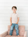 A young boy stands barefoot on a dusty pink tufted armchair against a white wood-paneled wall, wearing a short-sleeve button-up shirt with a tiny multicolor floral print and blue jeans.