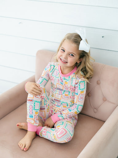 A young girl with a white bow in her hair sits on a dusty pink tufted armchair, wearing a long-sleeve pajama set with a colorful retro block pattern and pink trim.