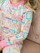A close-up of a child wearing a long-sleeve pajama top with a colorful shopping bag pattern on a white background and pink ribbed collar and cuffs, seated on a beige tufted chair.