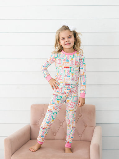 A young girl stands barefoot on a pink tufted chair against a white wood-paneled wall, wearing a long-sleeve pajama top and matching pajama pants with a colorful geometric pattern and pink trim.