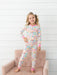 A young girl stands barefoot on a pink tufted chair against a white wood-paneled wall, wearing a long-sleeve pajama top and matching pajama pants with a colorful geometric pattern and pink trim.