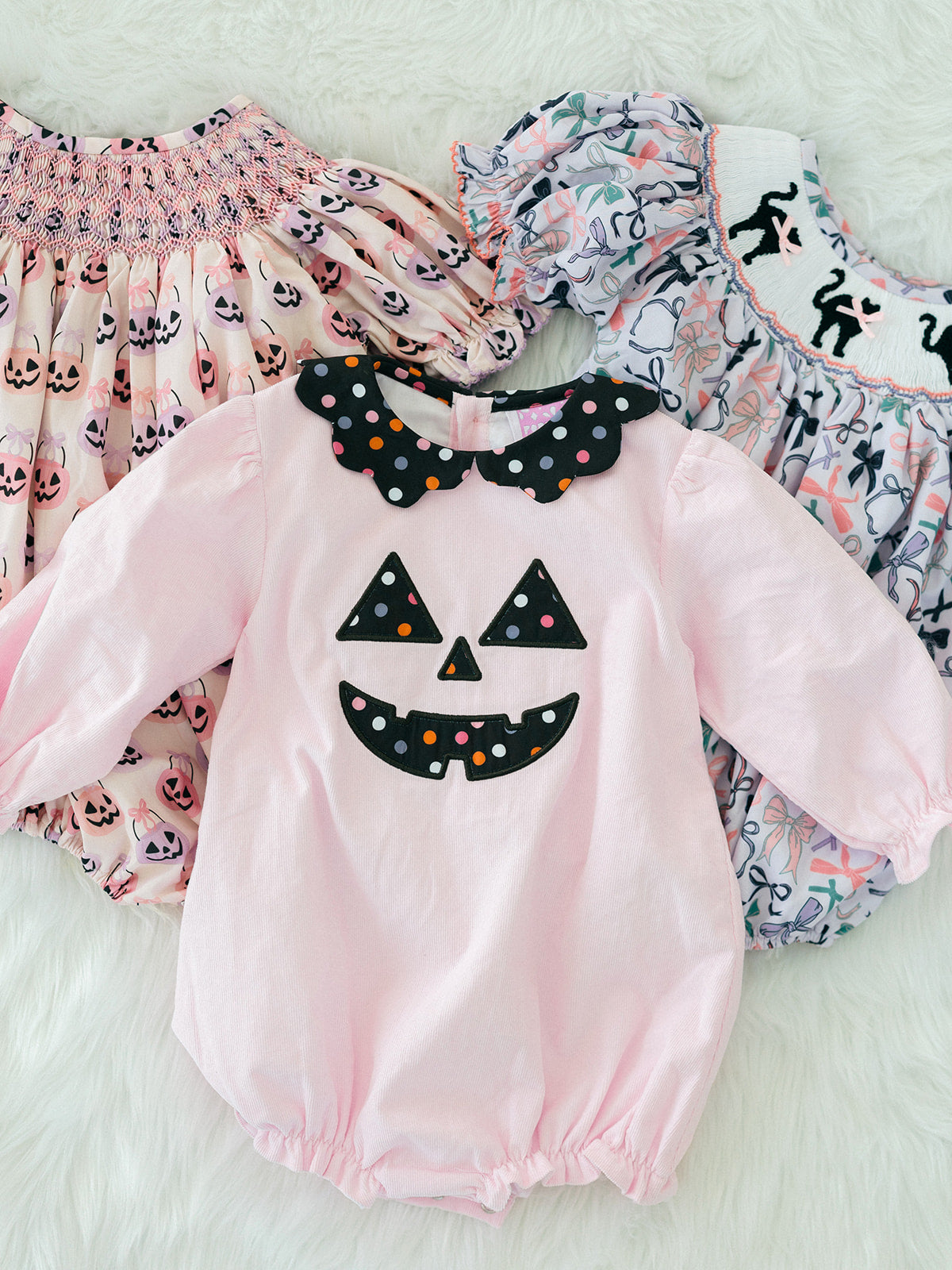 A baby long-sleeve romper in light pink with a black polka-dot jack-o'-lantern face applique on the chest and a matching black polka-dot bow collar. The romper is displayed flat on a white faux fur background, layered over two patterned baby smock dresses.