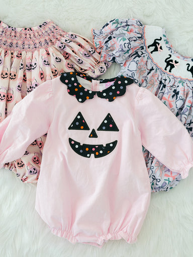 A baby long-sleeve romper in light pink with a black polka-dot jack-o'-lantern face applique on the chest and a matching black polka-dot bow collar. The romper is displayed flat on a white faux fur background, layered over two patterned baby smock dresses.