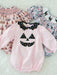 A baby long-sleeve romper in light pink with a black polka-dot jack-o'-lantern face applique on the chest and a matching black polka-dot bow collar. The romper is displayed flat on a white faux fur background, layered over two patterned baby smock dresses.