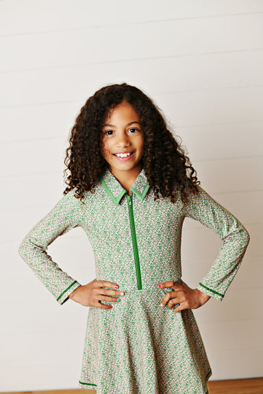 A young girl with curly brown hair smiling and posing with her hands on her hips, wearing a green and white patterned long-sleeve collared tennis dress with a green zipper.