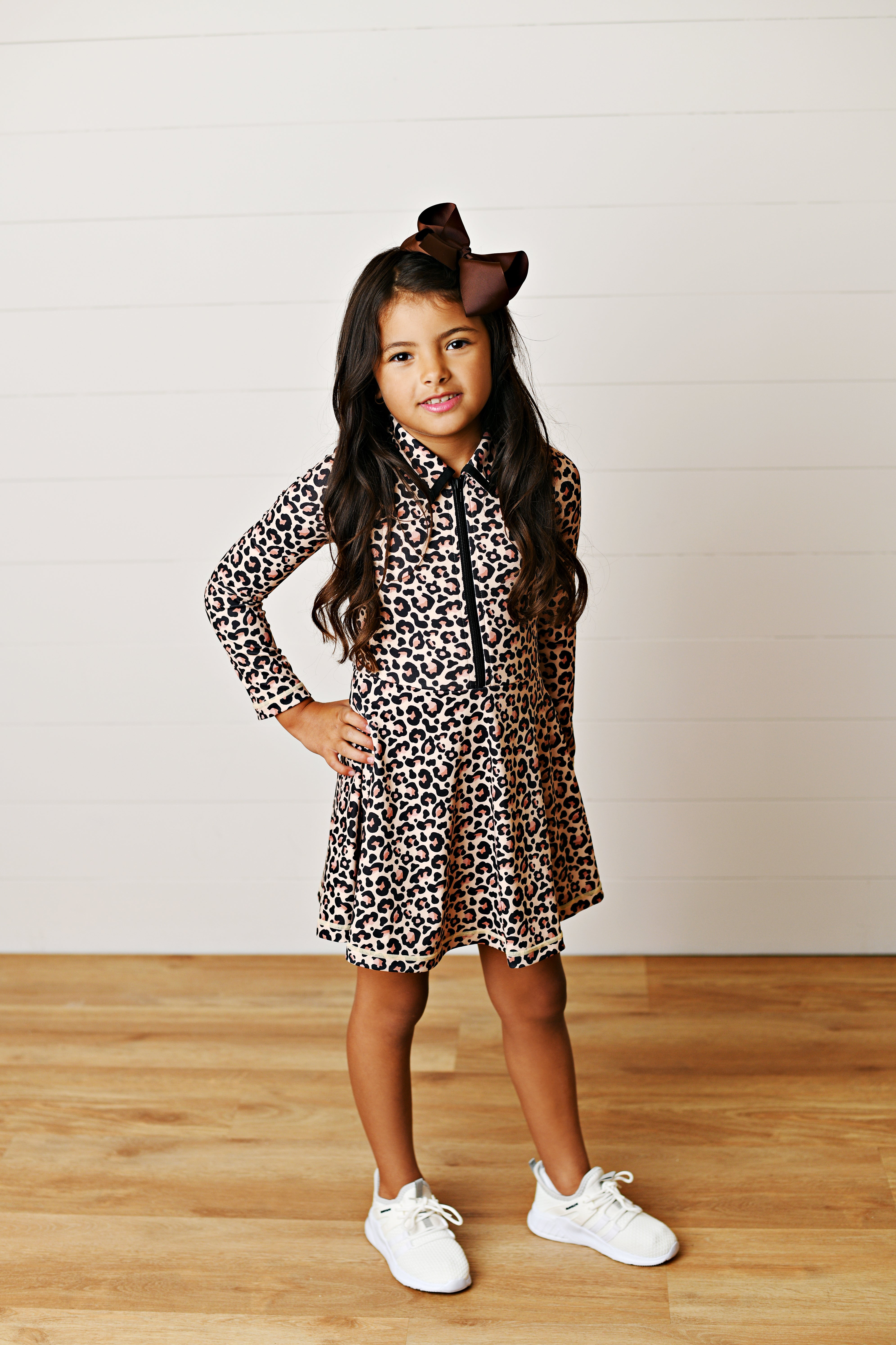 A young girl with long dark hair wearing a leopard-print long-sleeve collared tennis dress, a brown bow, and white sneakers, standing against a white shiplap wall and light wood floor.