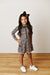 A young girl with long dark hair wearing a leopard-print long-sleeve collared tennis dress, a brown bow, and white sneakers, standing against a white shiplap wall and light wood floor.