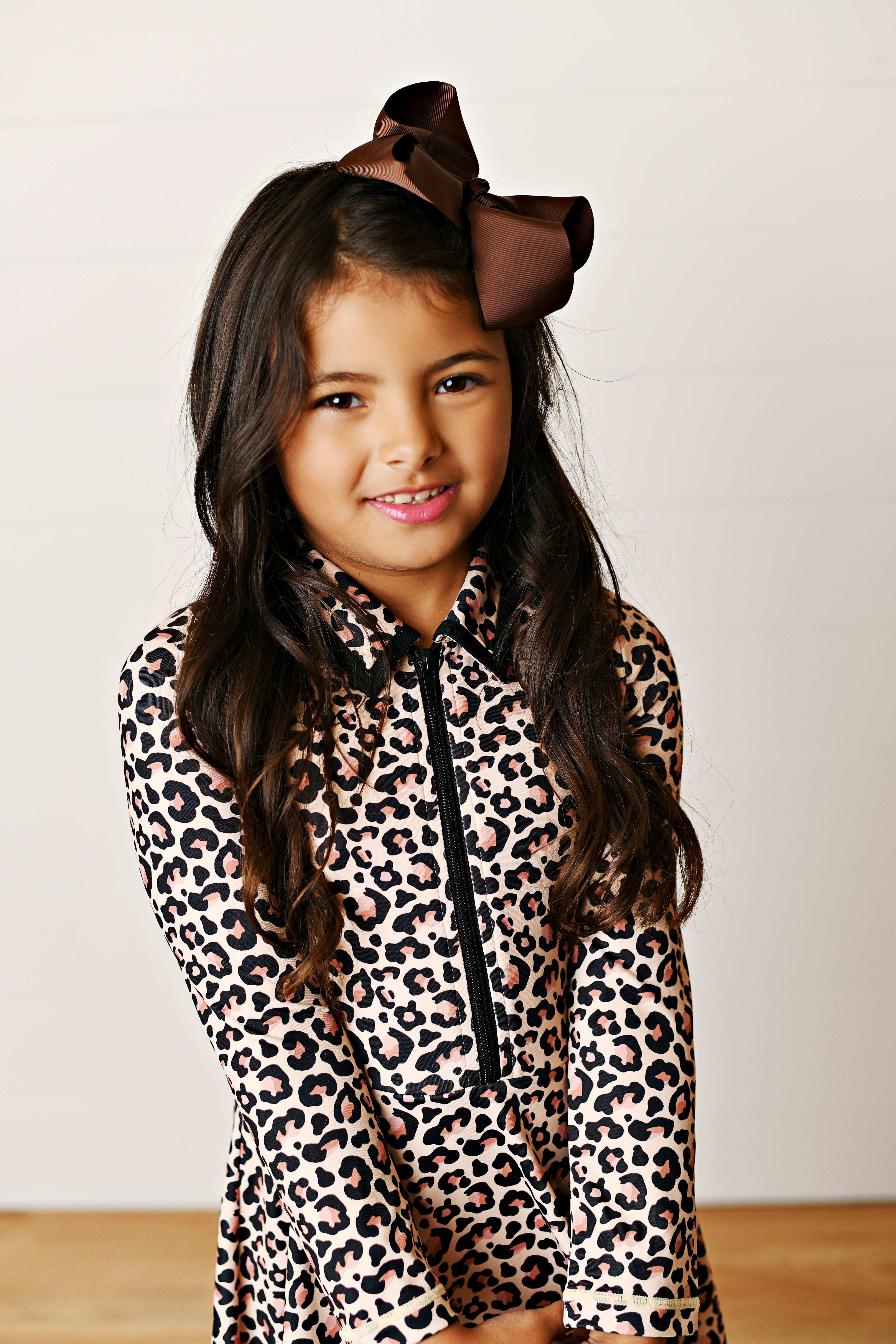 A young girl with long dark hair wearing a brown bow headband and a beige leopard print long-sleeve top with a black front zipper, posing against a plain off-white wall.
