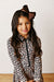 A young girl with long dark hair wearing a brown bow headband and a beige leopard print long-sleeve top with a black front zipper, posing against a plain off-white wall.