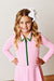 A young girl with blonde hair wearing a pink long-sleeve tennis dress with a green zipper and green collar trim, posing with hands on hips against a light beige wall.