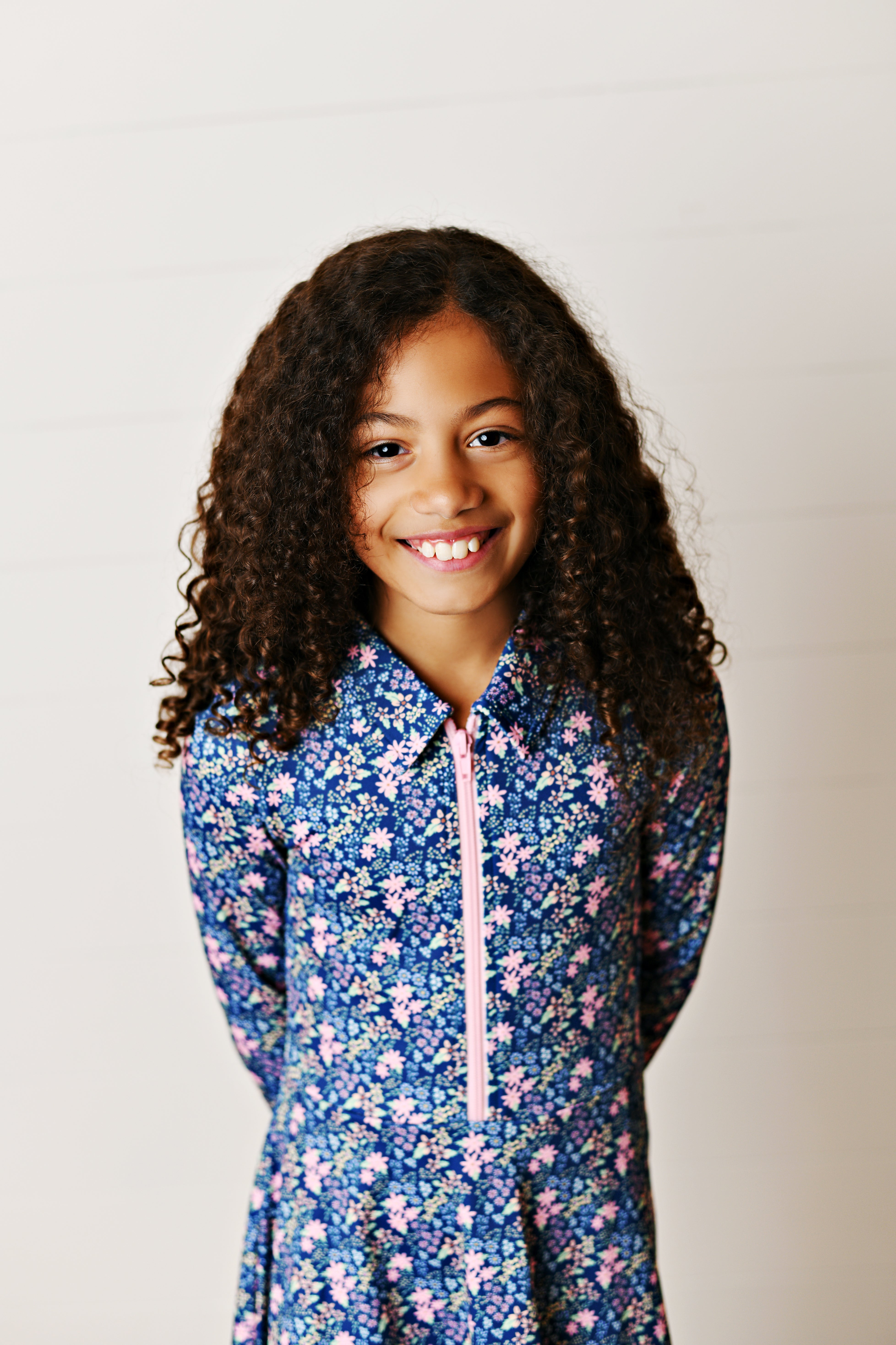 A smiling young girl with long curly brown hair wearing a blue long-sleeve collared tennis dress with a pink zipper and a pink, white, and blue floral pattern, set against a light off-white paneled wall.