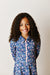 A smiling young girl with long curly brown hair wearing a blue long-sleeve collared tennis dress with a pink zipper and a pink, white, and blue floral pattern, set against a light off-white paneled wall.