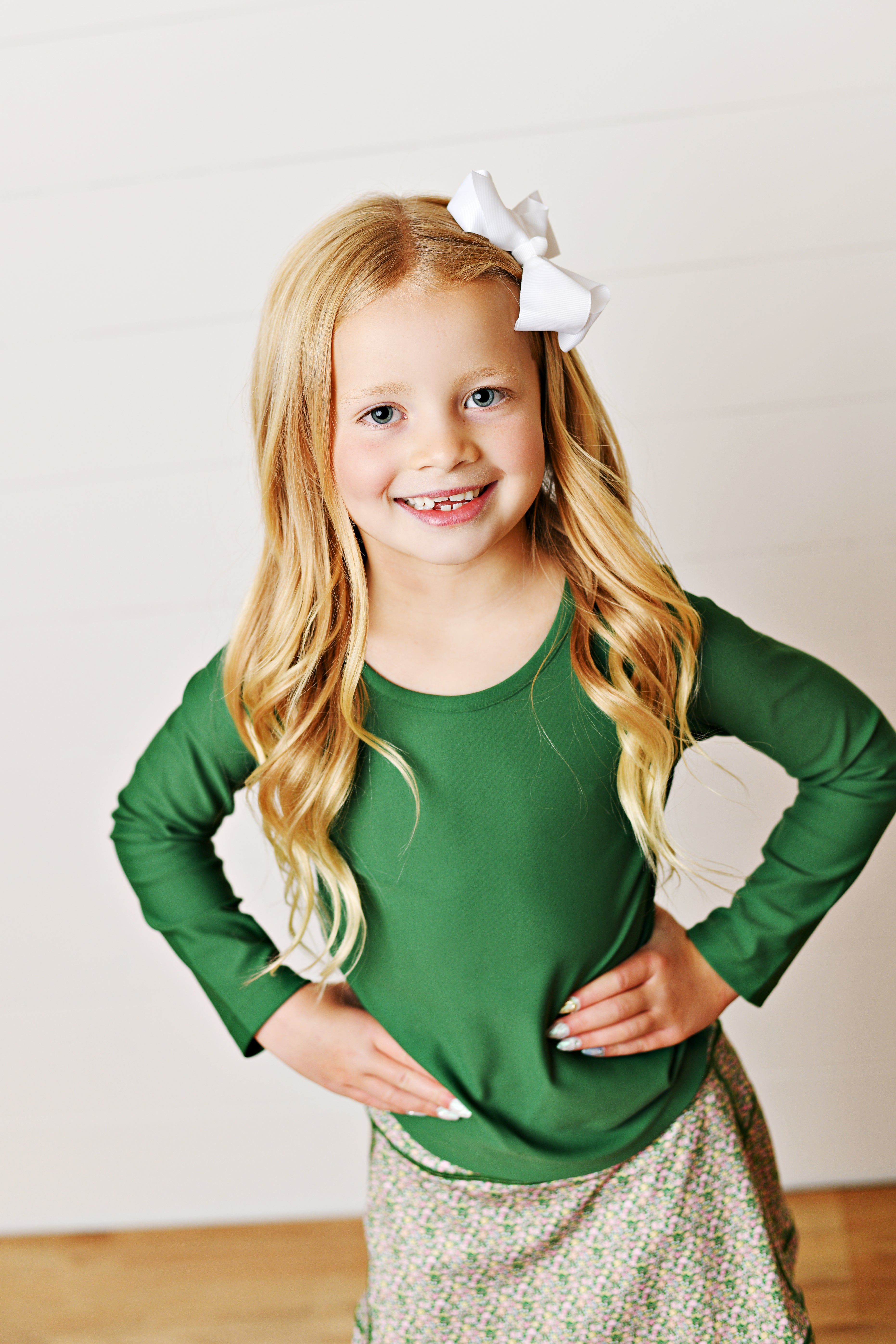 A young girl with long blonde hair wearing a green long-sleeve top and a patterned skirt, posing with hands on hips against a white paneled wall.