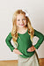 A young girl with long blonde hair wearing a green long-sleeve top and a patterned skirt, posing with hands on hips against a white paneled wall.