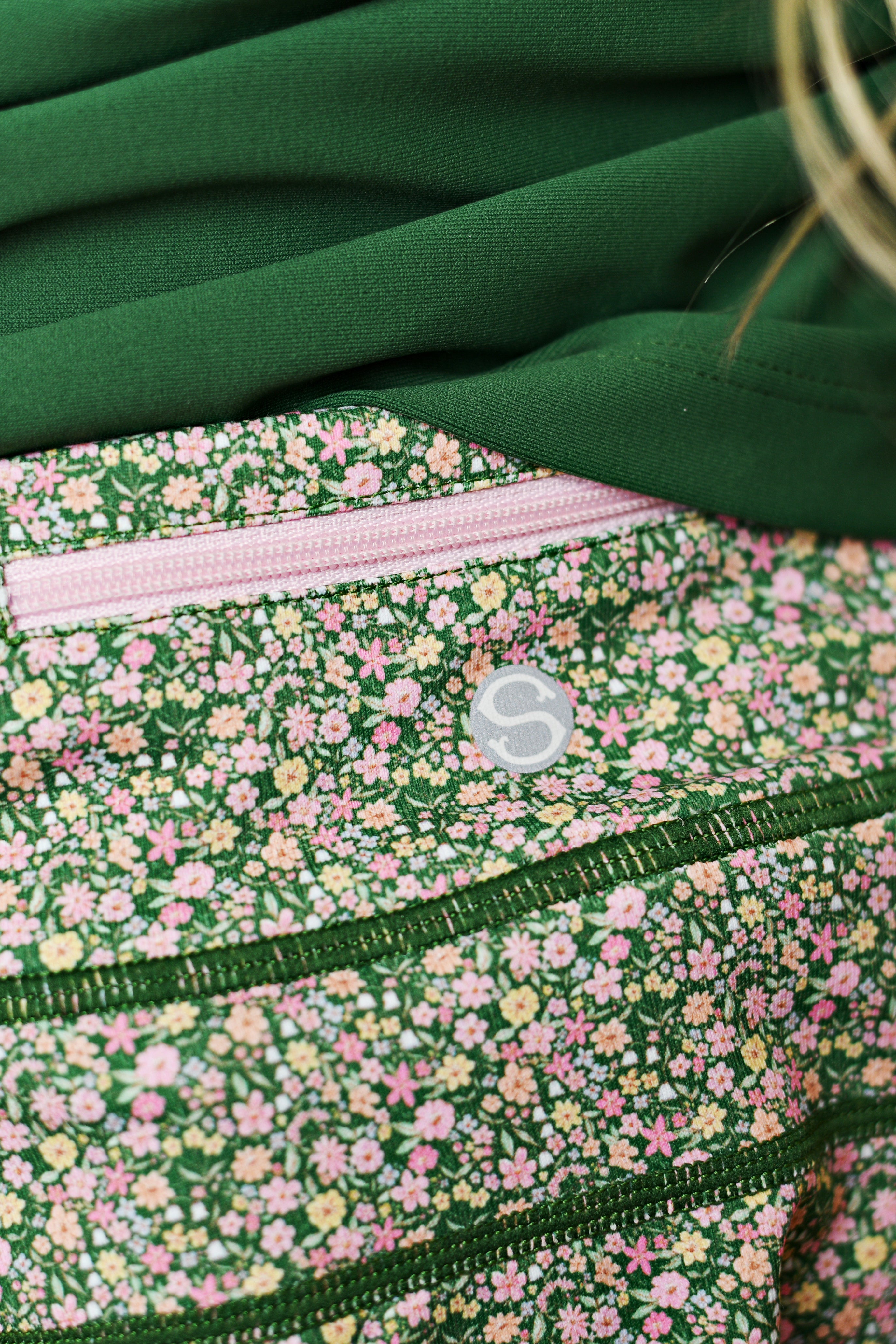 Close-up of a green floral-patterned fabric with a pink zipper pocket and a small white circular logo, alongside a solid dark green section.