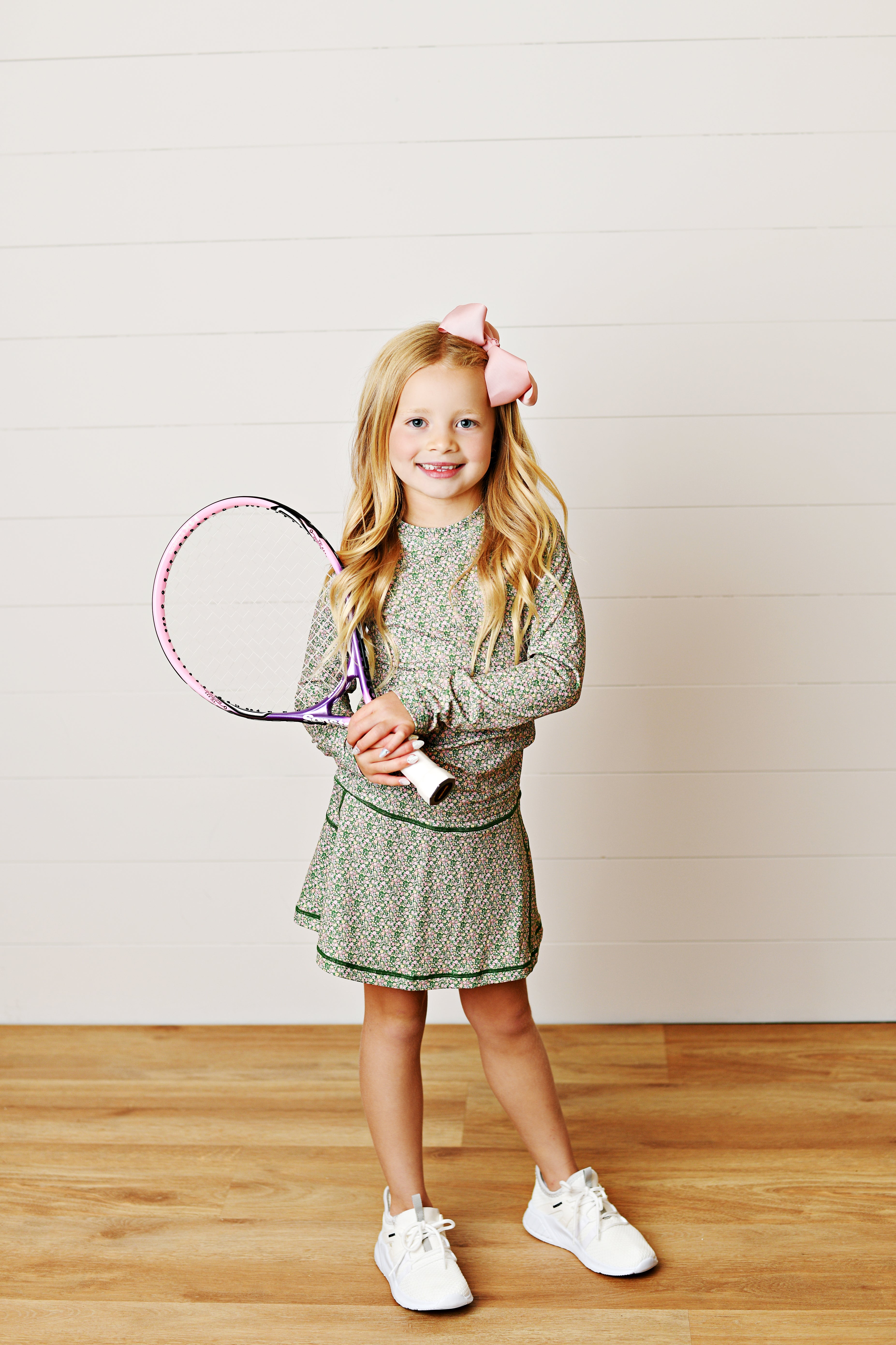 A young girl with blonde hair and a pink bow stands on a wooden floor against a white paneled wall, holding a tennis racket. She is wearing a green and white patterned long-sleeve tennis dress and white sneakers.