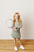 A young girl with blonde hair and a pink bow stands on a wooden floor against a white paneled wall, holding a tennis racket. She is wearing a green and white patterned long-sleeve tennis dress and white sneakers.