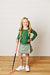 A young girl with long blonde hair stands indoors against a white paneled wall on a wood floor, holding a pink and black golf club. She is wearing a green long-sleeve top, a green and white patterned skirt, and white sneakers.