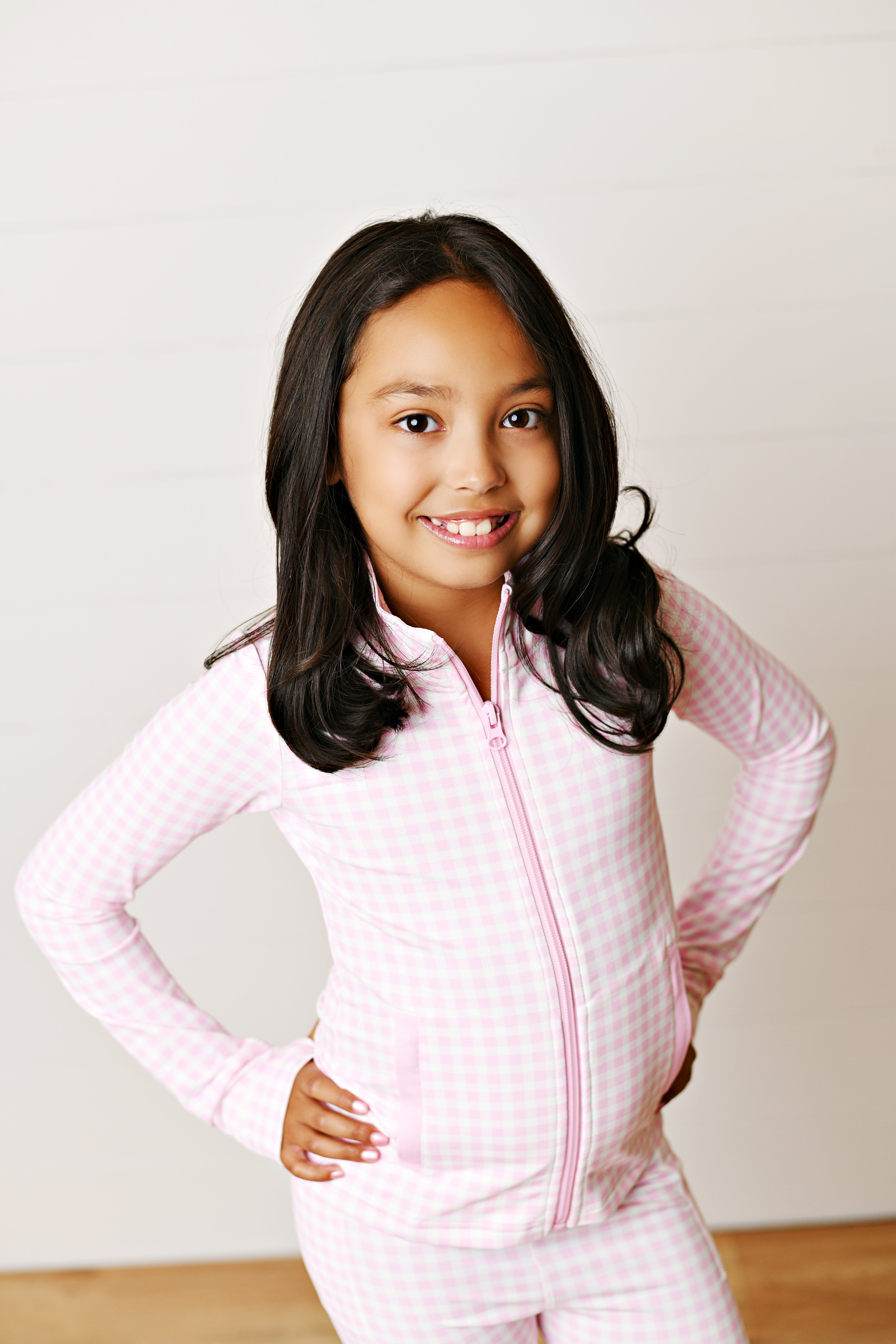 A young girl with long dark hair smiling and posing in a light pink and white gingham zip-up jacket and matching pants against a white paneled wall.