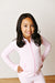 A young girl with long dark hair smiling and posing in a light pink and white gingham zip-up jacket and matching pants against a white paneled wall.